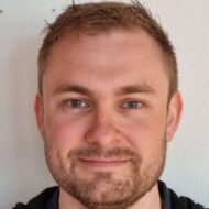 En mann med kort, lysebrunt hår, trimmet skjegg og blå øyne smiler lett mens han ser inn i kameraet. Bakgrunnen er en vanlig, lys vegg.