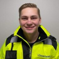 En ung person iført en gul jakke med ordet "Låssenteret" påtrykt, står og smiler foran en ensfarget bakgrunn.