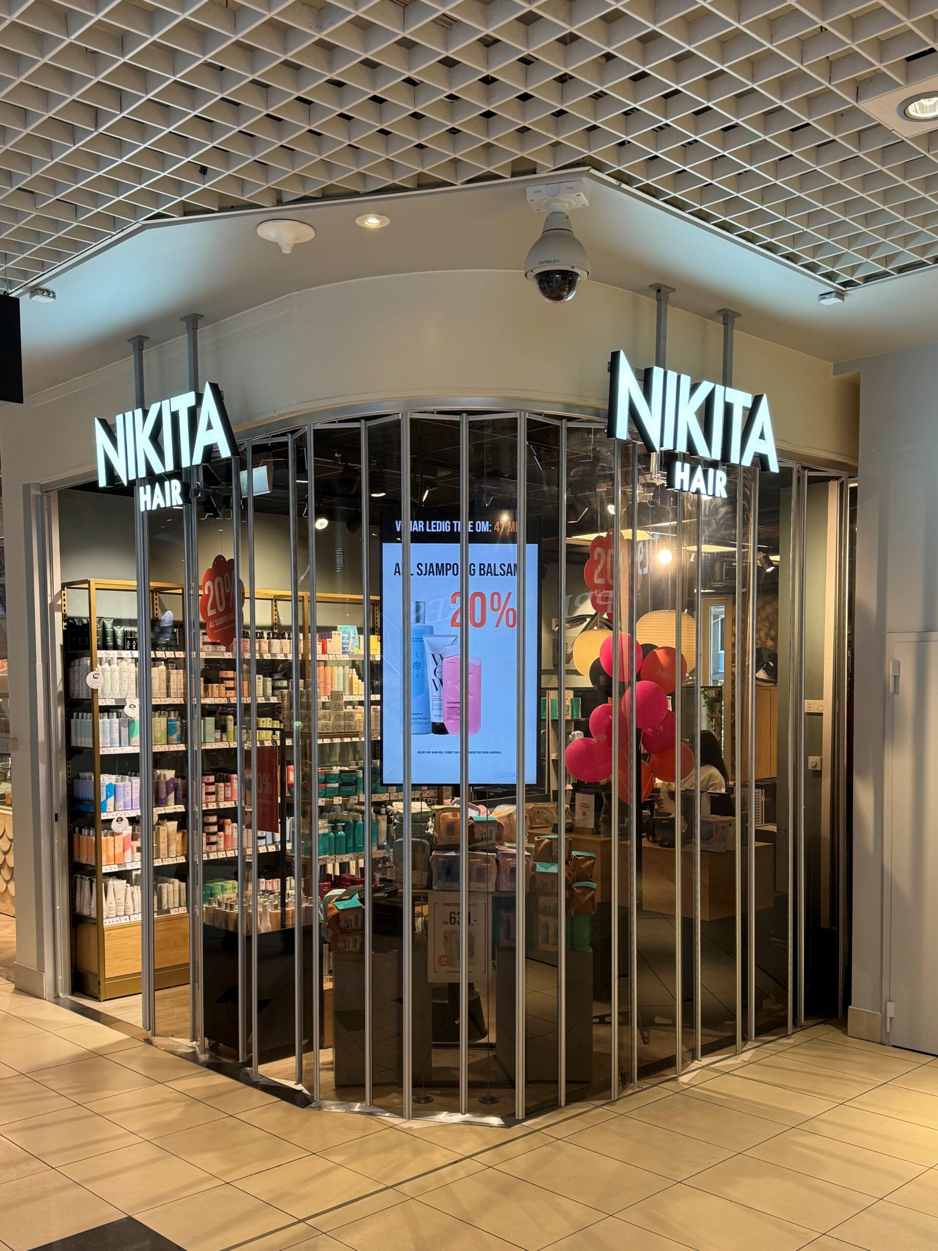 En Nikita Hair-frisørsalong med glassvegger, produktreoler, en digital skjerm som viser en rabatt og ballonger ved inngangen inne i et kjøpesenter.
