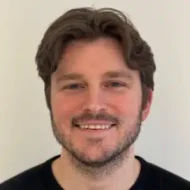 En mann med mellomlangt brunt hår og kort skjegg smiler til kameraet mot en ensfarget, lys bakgrunn.