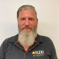 En mann med langt, grått skjegg og bakoverkjemmet hår, iført svart polo med "FLEXi lås og gitter"-logo, står mot en hvit, ensfarget vegg.