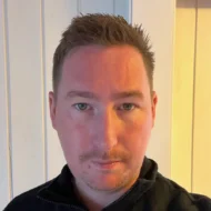 Alexander_mosaether En mann med kort, brunt hår og lyst ansiktshår, iført mørk jakke, står foran en hvit panelvegg og ser rett inn i kameraet.