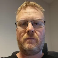 En mann med lyst hår, kort skjegg og briller ser direkte inn i kameraet i en innendørssituasjon.