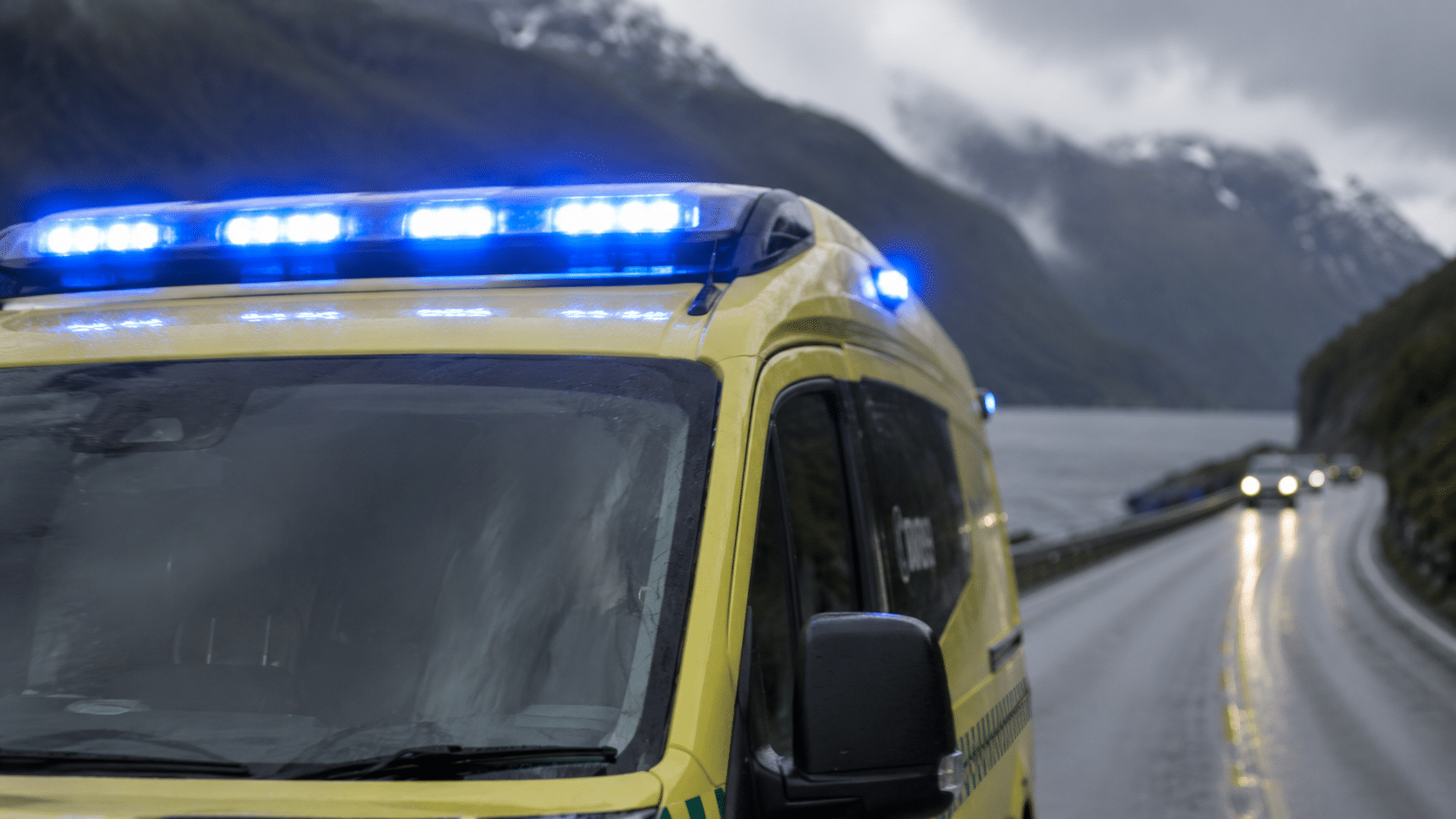 En gul ambulanse med blå blinkende lys står parkert på en våt, svingete vei ved siden av et vann og fjellene under en overskyet himmel.