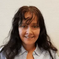 En kvinne med langt mørkt hår og lyseblå skjorte smiler til kameraet foran en hvit, strukturert vegg.