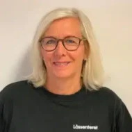 Anne-Wee-Førre-i-farger-scaled-e1597741872531-300x300 En kvinne med skulderlangt, blondt hår og briller, iført en svart skjorte med påskriften "Låssenteret", står mot en ensfarget, lys bakgrunn.