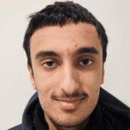 Ans-Iqbal-bilde-e1776691592505 En ung mann med kort, mørkt hår og ansiktsbehåring står vendt mot kamera, iført mørk jakke, mot en ensfarget, lys bakgrunn.