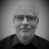 En middelaldrende mann med briller, kort, hvitt hår og skjegg, iført mørk skjorte med krage, står vendt mot kameraet med et nøytralt uttrykk. Bildet er i svart-hvitt.