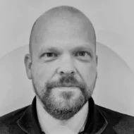 En skallet mann med skjegg og bart, iført mørk jakke, ser direkte inn i kameraet mot en ensfarget bakgrunn. Bildet er i svart-hvitt.