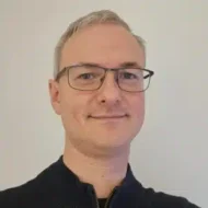 En person med kort, lyst hår og briller smiler lett, har på seg en mørk topp og står foran en vanlig, lys bakgrunn.