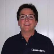 Berit-Drammen-e1599463450959 En person med briller og en marineblå skjorte med "Låssenteret" på står foran en hvit vegg og smiler til kameraet.