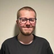 Bilde-Magnus-Sorvik-e1719820179683 En person med kort, blondt hår, briller og kort skjegg står opp mot en ensfarget, lys vegg, iført en mørk t-skjorte.
