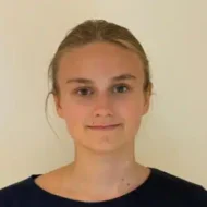 En person med lys hud og blondt hår som er trukket bakover, iført en mørk overdel, står vendt mot kameraet mot en vanlig lys bakgrunn.