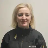 En kvinne med blondt hår og mørk jakke står mot en ensfarget, lys bakgrunn.