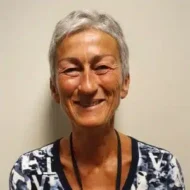 En eldre kvinne med kort, grått hår smiler til kameraet, iført mønstret skjorte og nøkkelbånd, foran en ensfarget, offwhite bakgrunn.