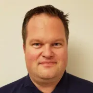 En mann med kort, brunt hår og lys hud står vendt mot kameraet og smiler lett, iført mørk skjorte, mot en ensfarget, lys bakgrunn.