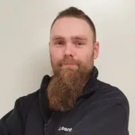 En mann med langt, fyldig skjegg og kort hår står mot en ensfarget, lys bakgrunn, iført en mørk jakke med "iSentio" synlig på brystet.