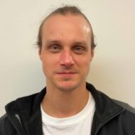 En mann med lys hud og blå øyne står mot en hvit vegg, iført en svart jakke over en hvit skjorte. Han har lysebrunt hår som er trukket bakover.