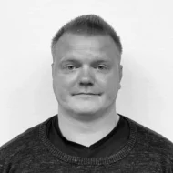 En mann med kort, lyst hår og nøytralt ansiktsuttrykk, iført en mørk genser over en mørk skjorte, står mot en ensfarget hvit bakgrunn.