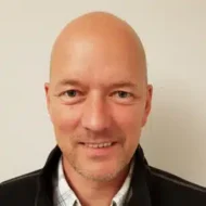En skallet mann med lys hud, iført mørk jakke over en rutete skjorte, smiler til kameraet mot en ensfarget, lys bakgrunn.