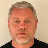 En middelaldrende mann med kort, grått hår, lyse øyne og skjegg ser direkte inn i kameraet, iført svart skjorte mot en ensfarget bakgrunn.