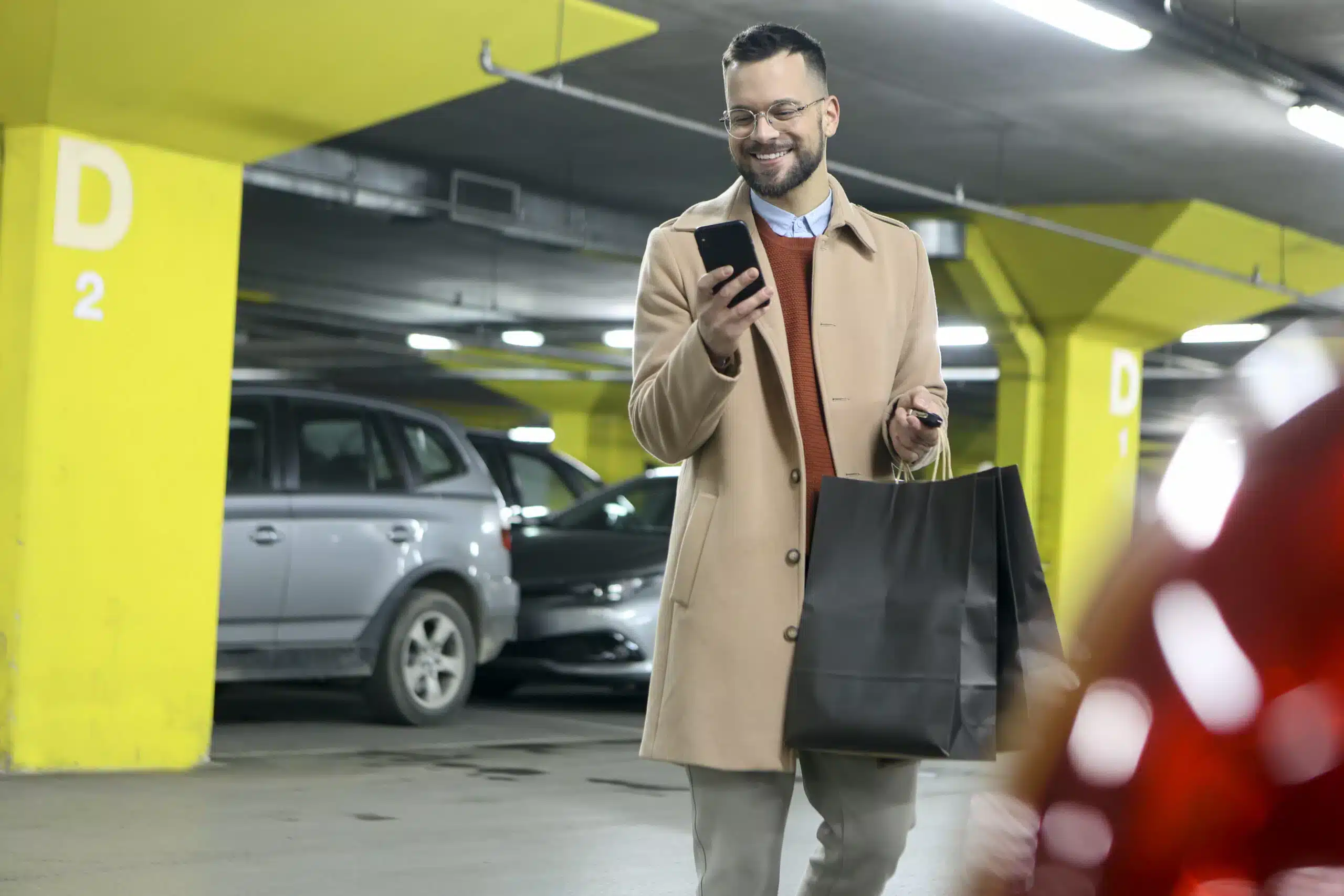 En mann i brun frakk smiler mens han ser på telefonen, med handleposer og bilnøkler i hånden, på vei inn i et innendørs parkeringshus.