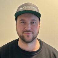 Gjovik-Lars-e1758201187832-300x300 En mann med kort skjegg, mørk caps med påskriften "IDT SPORTS" og svart skjorte står foran en ensfarget, lys vegg.