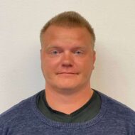 En mann med kort, blondt hår og mørk genser over en svart skjorte står mot en hvit vegg og ser direkte inn i kameraet med et nøytralt ansiktsuttrykk.