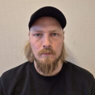 Hans-Erik-Omberg-scaled-e1745912236879-350x350 En mann med langt blondt skjegg og bart, iført svart caps og svart skjorte, ser direkte inn i kameraet mot en ensfarget, lys bakgrunn.