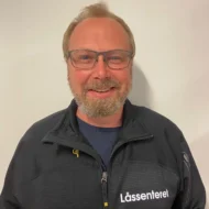 Hans-Petter-Korsane-rotated-e1599549935830 En mann med briller og skjegg, iført en svart jakke med påskriften "Låssenteret", står foran en ensfarget, lys vegg.