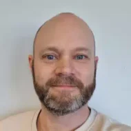 En mann med skallet hode, lys hud, blå øyne og skjegg ser inn i kameraet og smiler lett mot en ensfarget, lys bakgrunn.