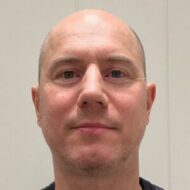 En middelaldrende mann med barbert hode og lyst ansiktshår ser direkte inn i kameraet mot en ensfarget, lys bakgrunn.