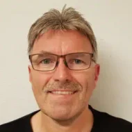 En mann med briller og bart smiler til kameraet.