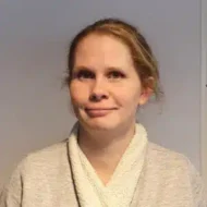Henriett_jellebo-300x300 En kvinne med lys hud og rødblondt hår, iført en lysegrå genser over en hvit topp, står mot en ensfarget bakgrunn og ser litt til siden.