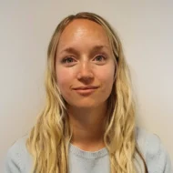 En kvinne med langt blondt hår og en lyseblå genser ser inn i kameraet mot en vanlig lys bakgrunn.