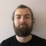 En person med helskjegg og bart, langt hår som er trukket bakover, og iført svart skjorte står mot en ensfarget, lys bakgrunn.