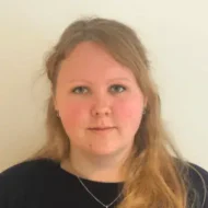 En person med langt blondt hår, lys hud og blå øyne står vendt mot kameraet mot en ensfarget, lys bakgrunn.