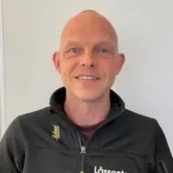 Jimmy_Helgesen-300x300 En skallet mann i svart jakke med glidelås og teksten "Låscenter" står foran en ensfarget, lys bakgrunn og smiler til kameraet.
