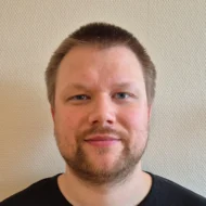 Joakim-Sanna En mann med kort, brunt hår, trimmet skjegg og svart skjorte står foran en strukturert, lys vegg og ser rett inn i kameraet.