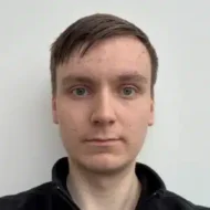 En ung person med kort, brunt hår og grønne øyne ser rett inn i kameraet mot en enkel, hvit bakgrunn, iført en svart topp med glidelås.