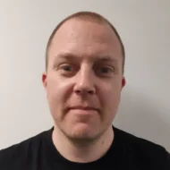 En mann med kort, lysebrunt hår og svart skjorte står vendt mot kameraet, mot en ensfarget, lys bakgrunn.