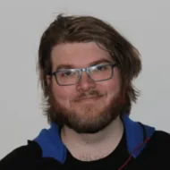 En person med briller, skjegg og mellomlangt brunt hår står vendt mot kameraet og smiler lett. Vedkommende har på seg en mørk topp med blå hette. Bakgrunnen er ensfarget og lys.