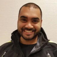 En mann med kort hår og skjegg smiler til kameraet, iført svart jakke, foran en hvit, strukturert vegg.