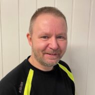 En mann med kort, lyst hår og skjegg, iført svart skjorte med gule aksenter, smiler foran en vegg med hvitt panel.