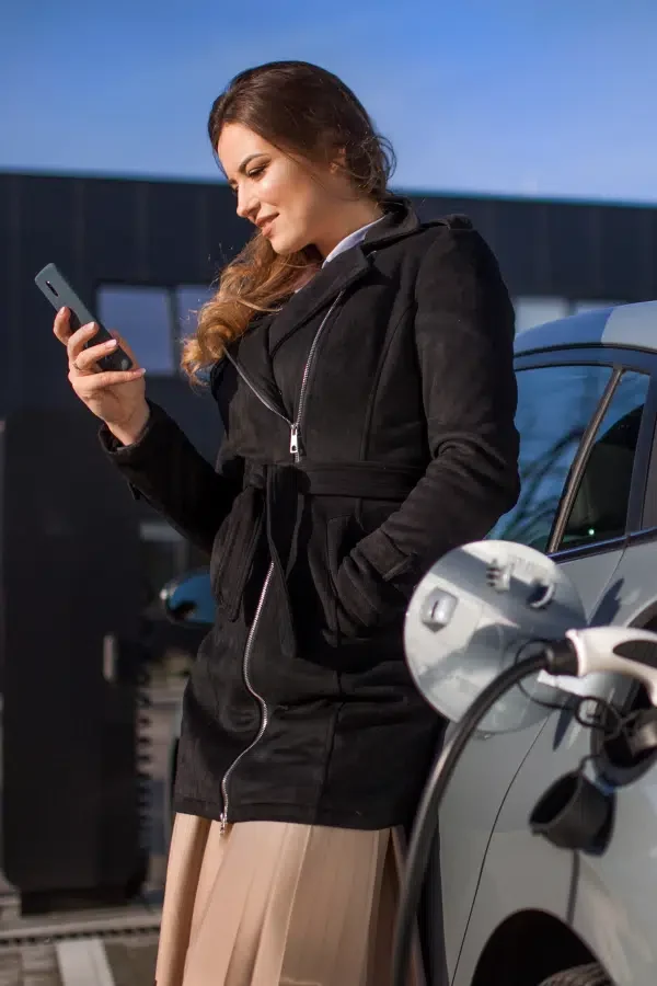 Kvinne i svart frakk står ved en elbil og ser på telefonen sin mens bilen lades på en utendørs ladestasjon.