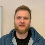 Magnus En mann med kort, blondt skjegg og bart, lys hud og kort hår har på seg en blå hettegenser over en svart skjorte og står foran en vegg med beige tekstur.