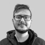 En person med kort hår, briller og skjegg står vendt mot kameraet, iført en mørk hettegenser mot en ensfarget, lys bakgrunn.