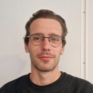 Mathias Nilsen En mann med kort brunt hår, briller og en svart skjorte står foran en helt hvit vegg og ser inn i kameraet med et nøytralt uttrykk.