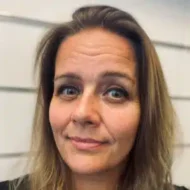 En kvinne med langt, lysebrunt hår og lyse øyne ser inn i kameraet med et nøytralt uttrykk. Hvite, horisontale veggpaneler er i bakgrunnen.