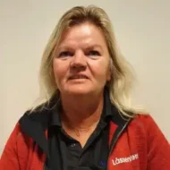 En kvinne med blondt hår og rød jakke over en svart skjorte står foran en ensfarget, lys bakgrunn.